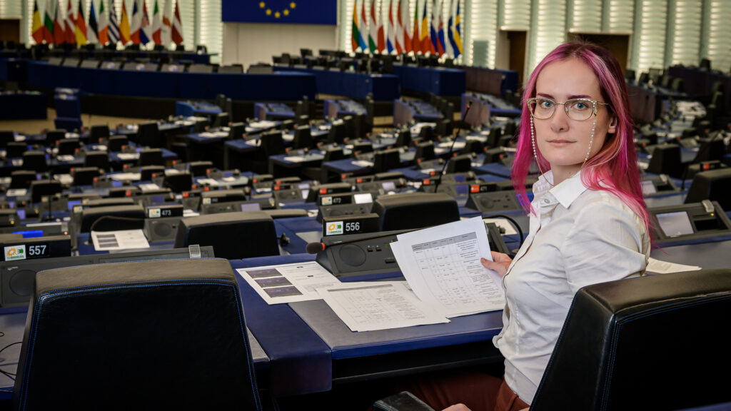 Markéta seating at the back of the empty plenary room, looking back at the camera
